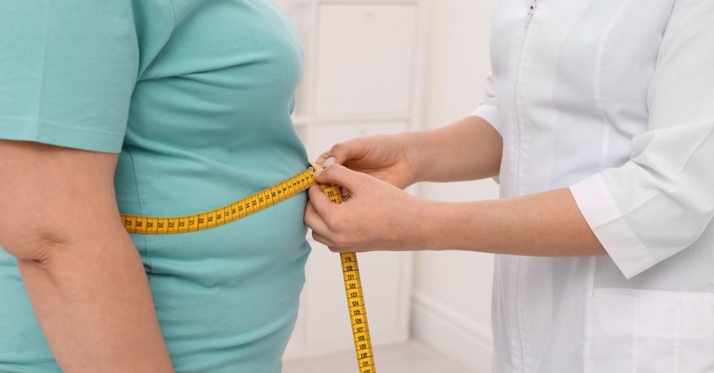 A medical professional, wearing a white coat, is using a yellow measuring tape to measure the waist circumference of a person in a light blue t-shirt. 