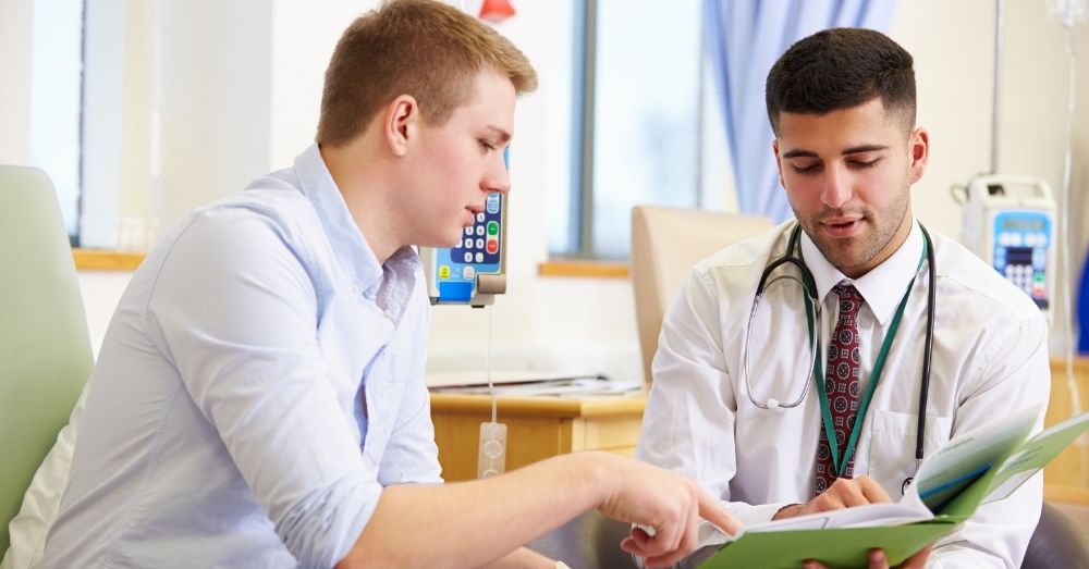 A male doctor with a stethoscope around his neck is seated and pointing at a document in a green folder he's holding.