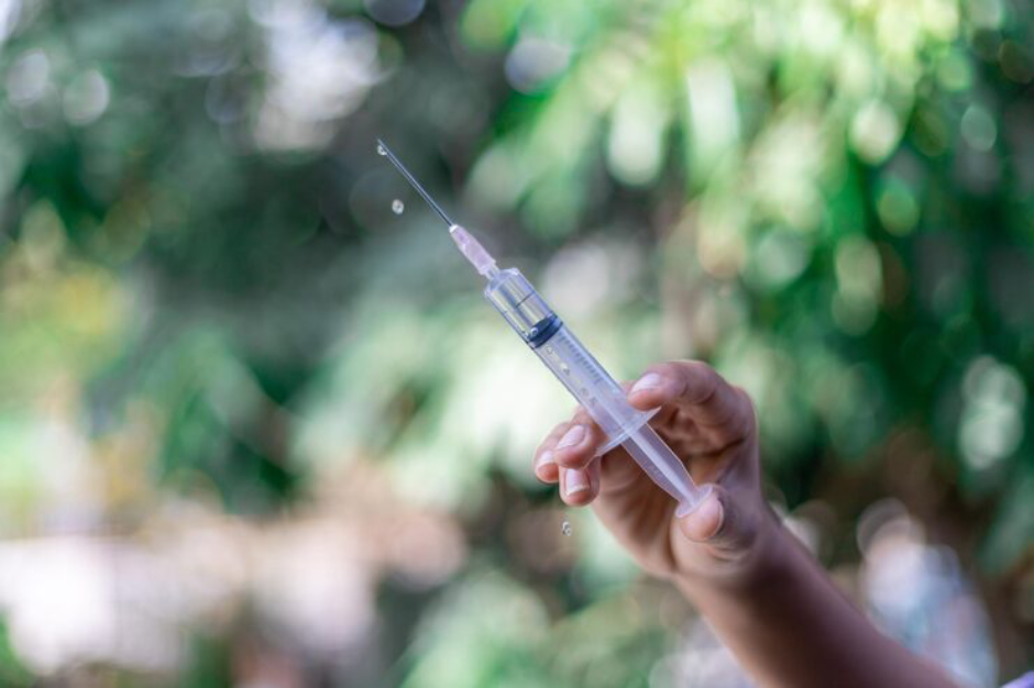 A close-up shows a hand holding a syringe with a clear liquid, from which a drop is falling from the needle tip against a blurred green, leafy background.