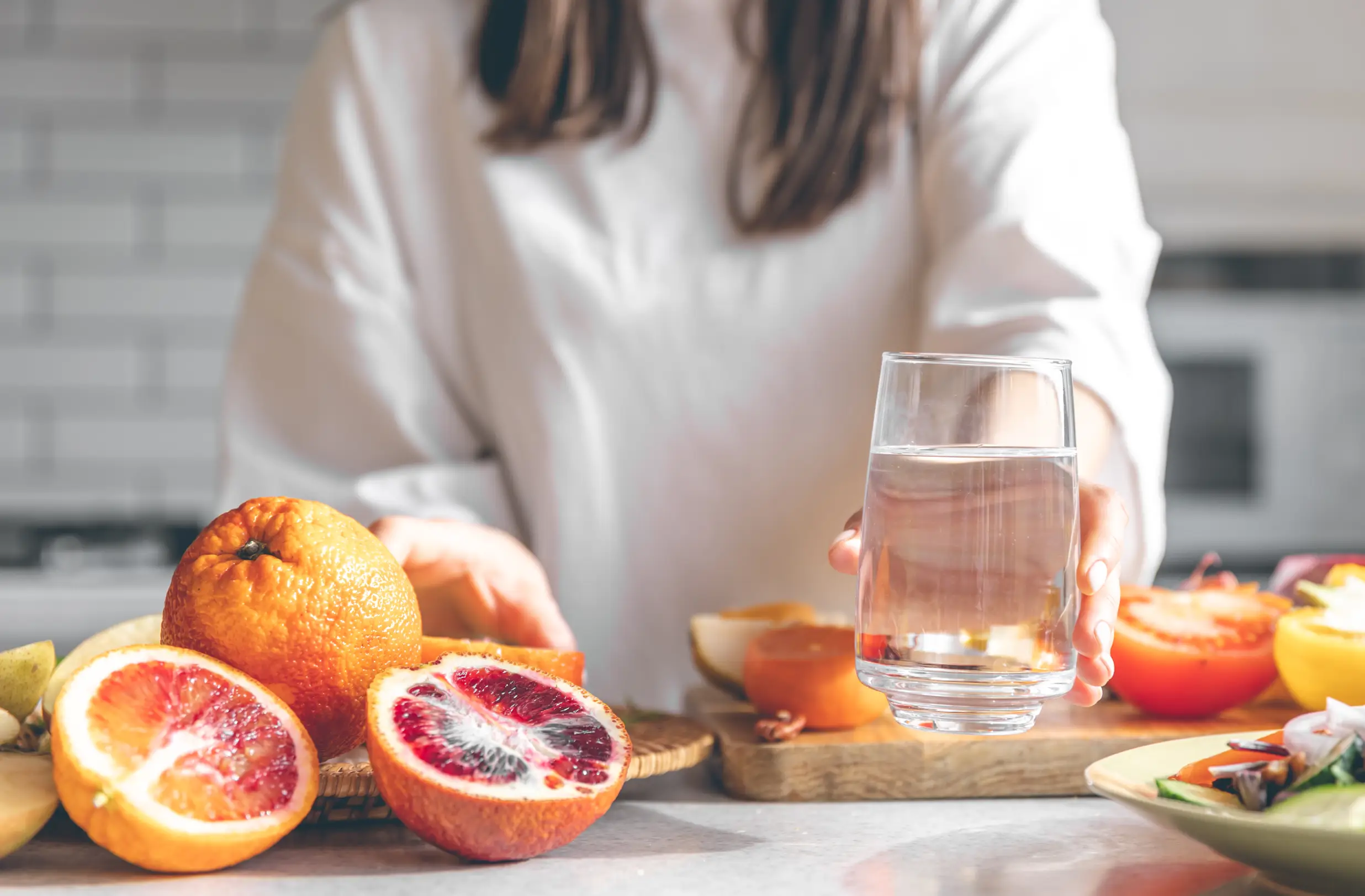 Woman holding a glass of water beside fresh citrus fruits and vegetables on a kitchen counter