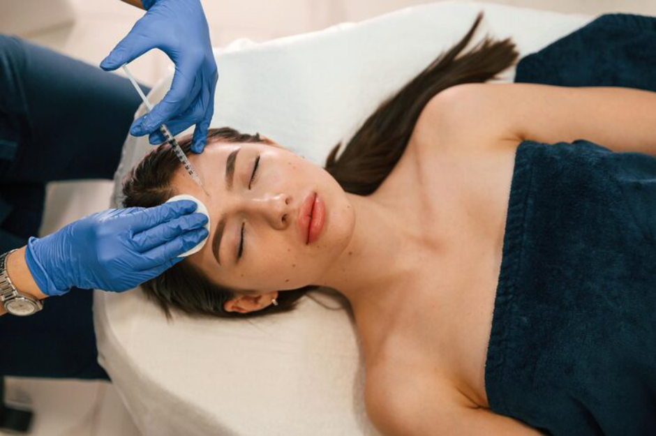 Beautician administering forehead botox injection to woman lying on treatment bed covered with dark towel.