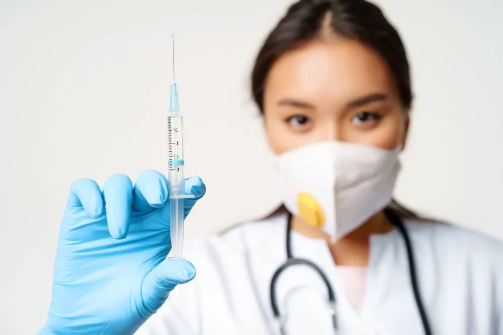 Doctor wearing protective mask and blue gloves holding syringe with needle in medical clinic.