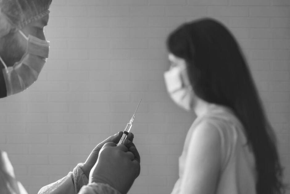 A black and white close-up shows a medical professional in a cap and mask holding a syringe with the needle pointing towards a blurred person in a mask in the background.