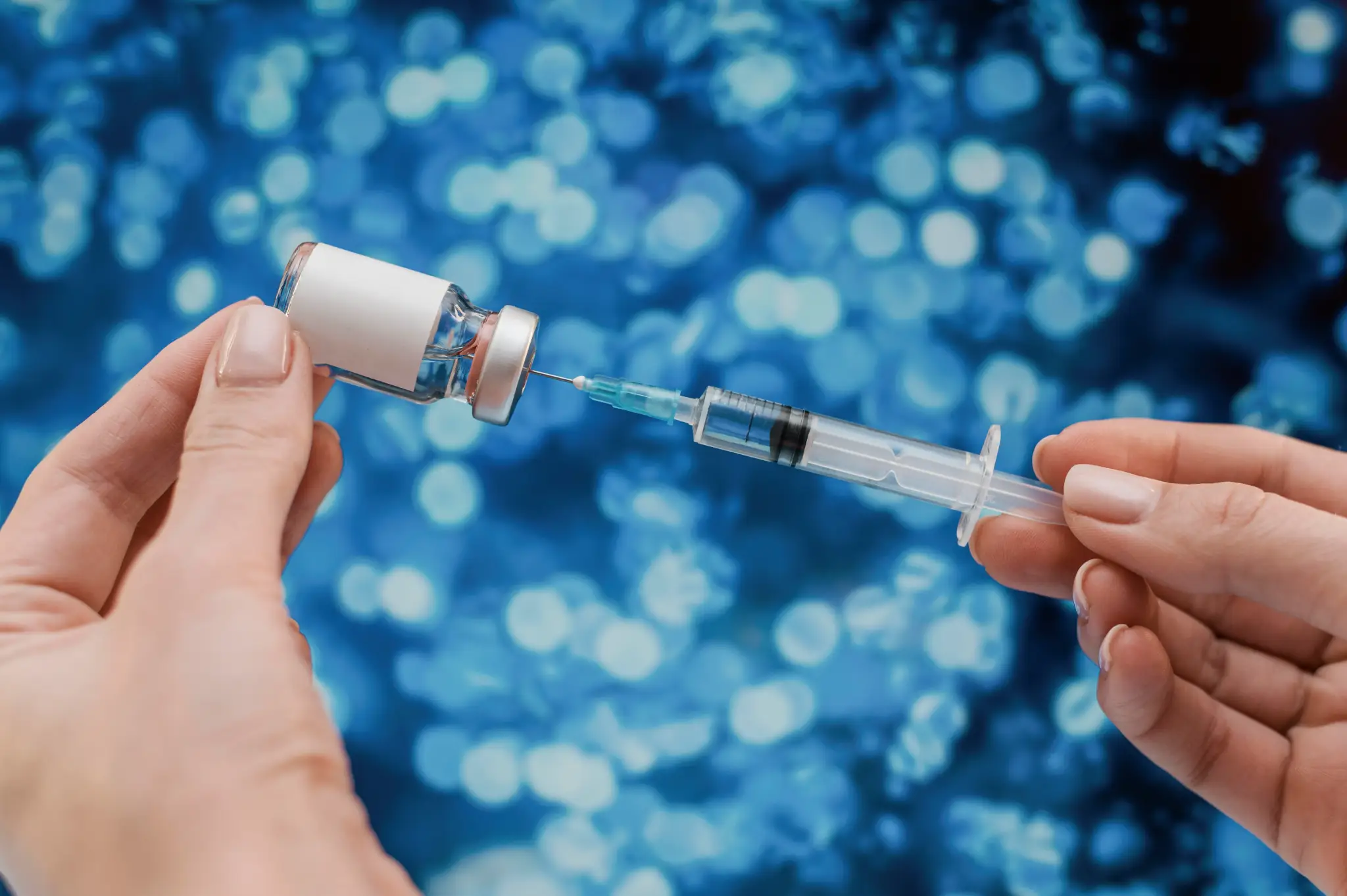 Close-up of hands drawing liquid from medical vial into syringe against blue bokeh background.