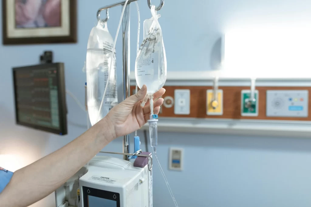 Close-up of nurse adjusting IV fluid bag in hospital room with medical equipment and monitor in the background