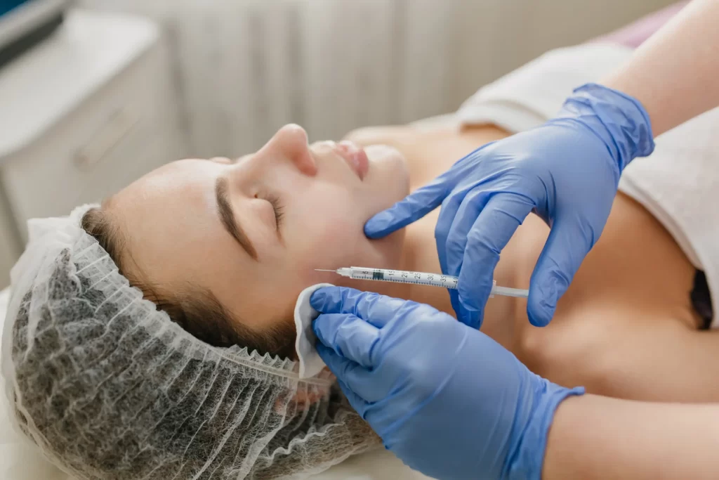 Cosmetologist performing cheek filler injection on woman lying with hair cap in aesthetic clinic.