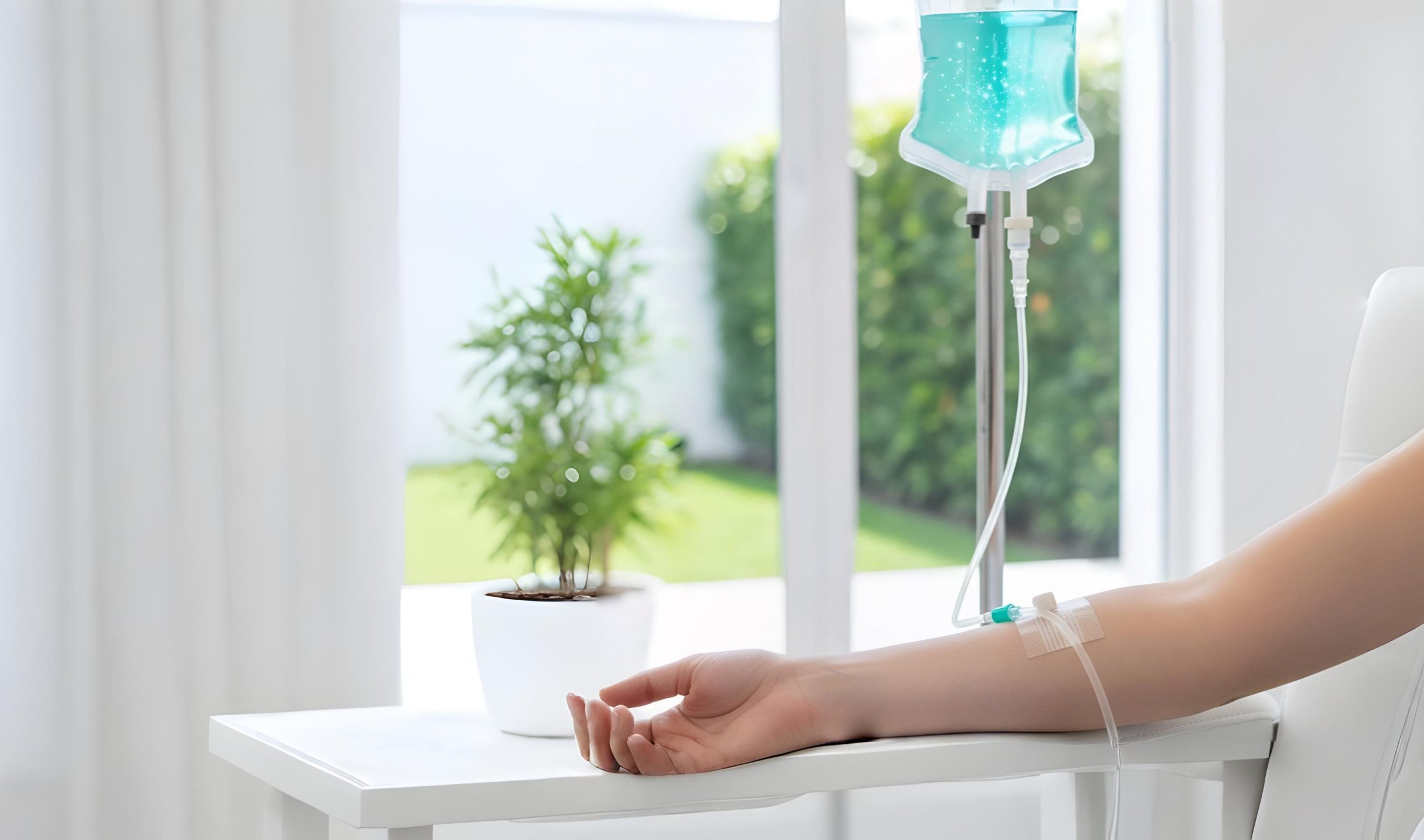 A close-up view of a patient’s arm resting on a white chair while receiving an IV infusion. A clear tube leads from a medical port on the forearm up toward a hanging IV bag filled with teal-colored fluid. The background is a bright, airy room with soft-focus greenery visible through a window.