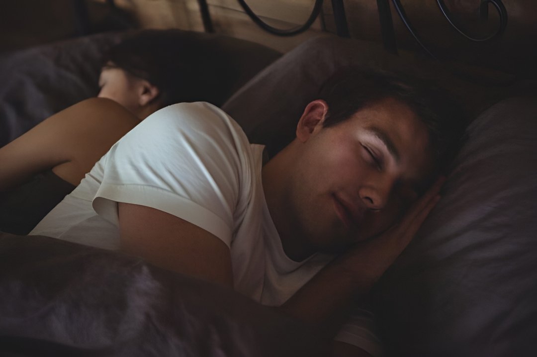 A sleeping man stretching and smiling in bed under a blanket in a bright, cozy bedroom during the morning.