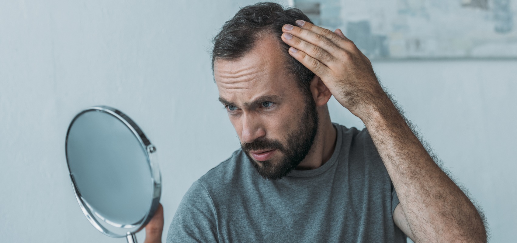 worried-looking man looks in a hand mirror while pulling back his hair