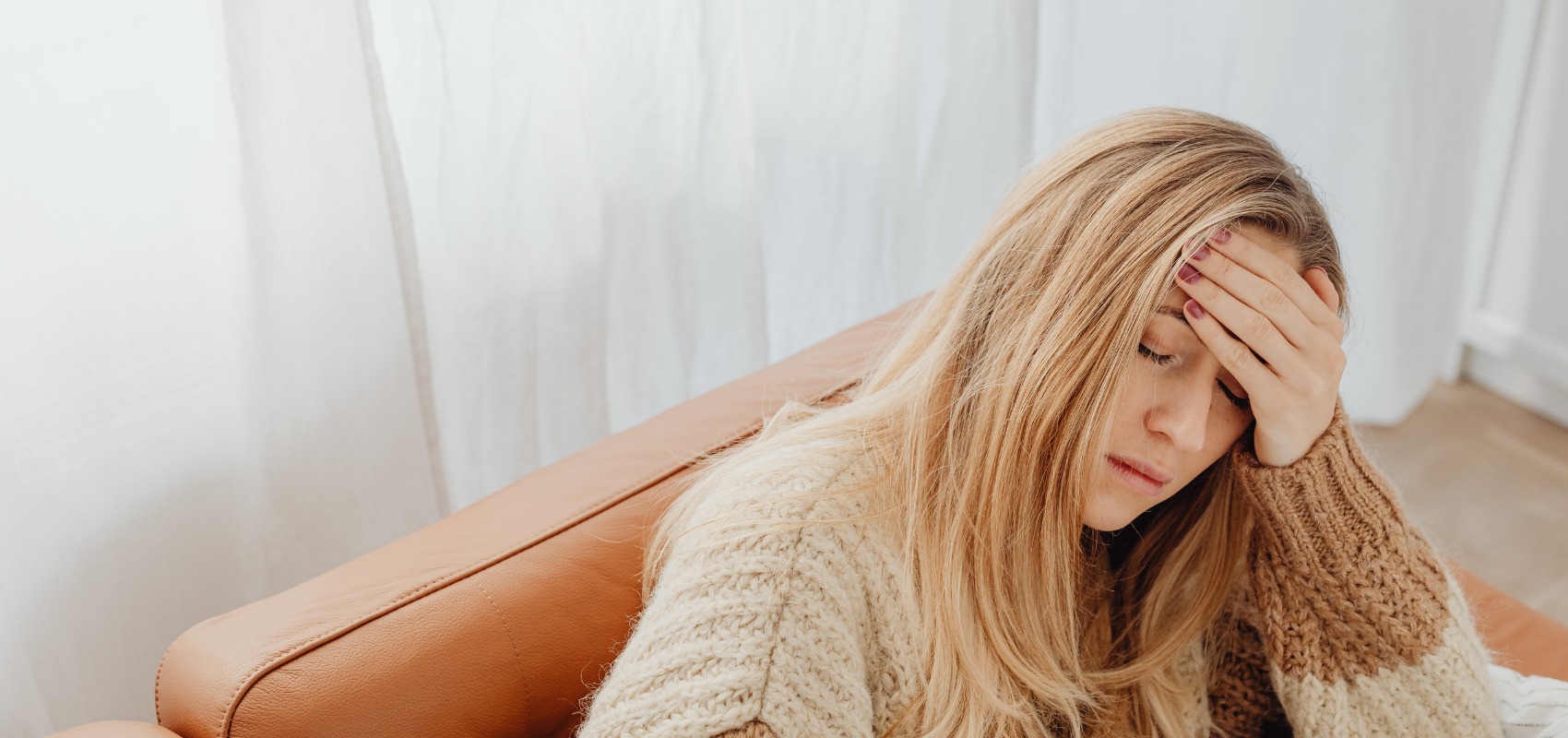 a woman sits on a brown leather couch, holding her hand to her forehead in stress