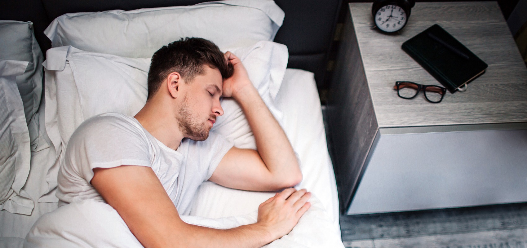 A man sleeps in a bed next to a nightstand with a clock