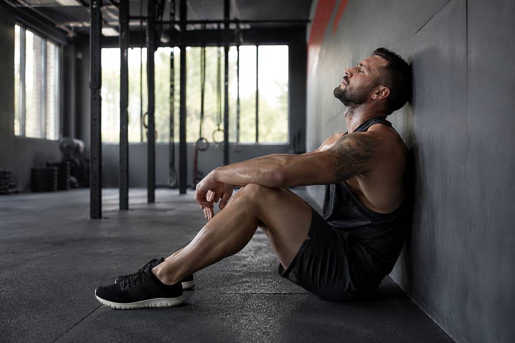 A male athlete sitting on a gym floor post-workout, eyes closed and head resting against a wall, cooling down after an intense session.